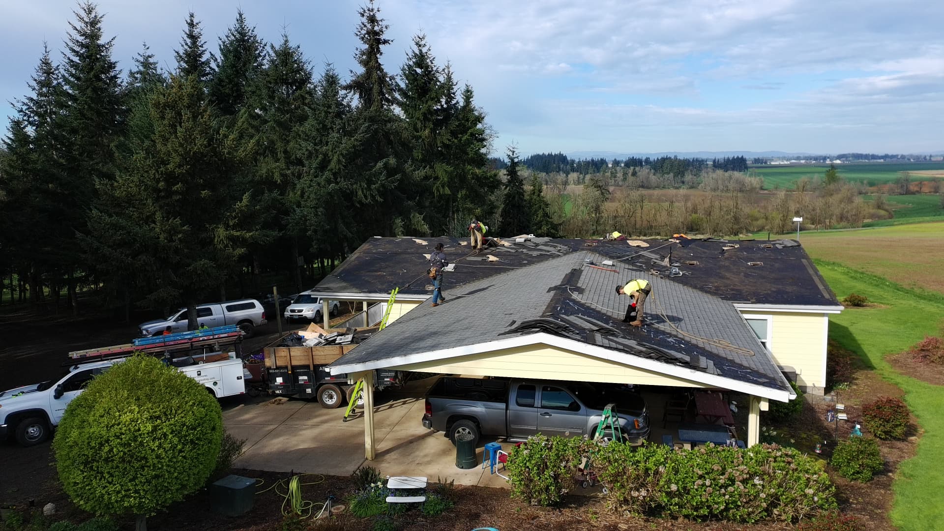Abiqua Roofing crew tearing off and re-roofing a single-story home surrounded by fir trees and farm fields